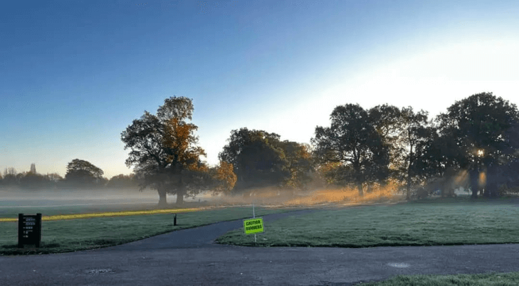 A sign reading CAUTION RUNNERS set up at a Parkrun event, with neatly mowed lawn and pretty trees in the background.