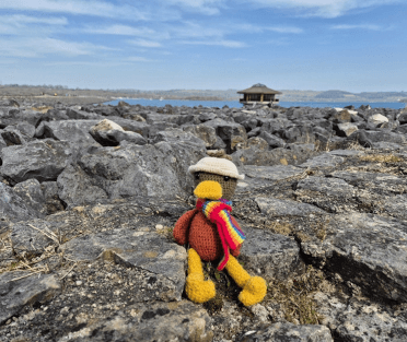 The Contours mascot, Croutons, a crocheted duck in a crocheted hat, sits on the rocky shore of Carsington Water with the reservoir behind him.