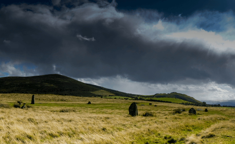 Dark clouds hang broodingly low over a wide open field studded with the standing stones at Mitchell's Fold, with impressive hilly backdrop.