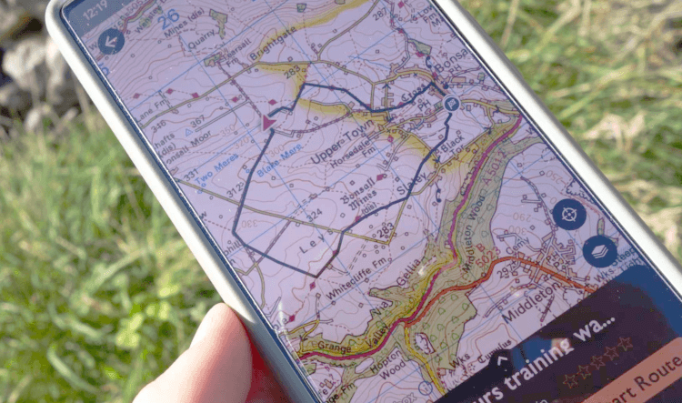A close-up of a phone, as held out on a walking trail. The screen displays the OS Map interface, loaded with a GPX file showing the route of one of Contours Holidays' circular training walks in the Peak District. The route is clearly displayed over Ordnance Survey mapping.